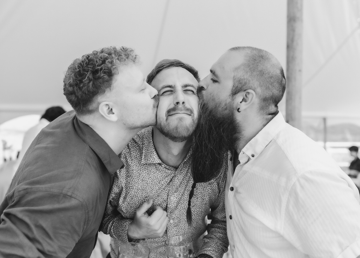 Three men pose playfully; the man in the center smiles with his eyes closed as the two others kiss his cheeks. All appear happy in a casual, candid moment under a tent.