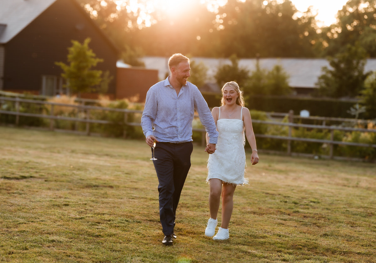 A man and woman, both smiling, walk hand in hand across a grassy field at sunset. The woman wears a white dress and sneakers; the man holds a drink. A wooden fence and buildings are in the background.