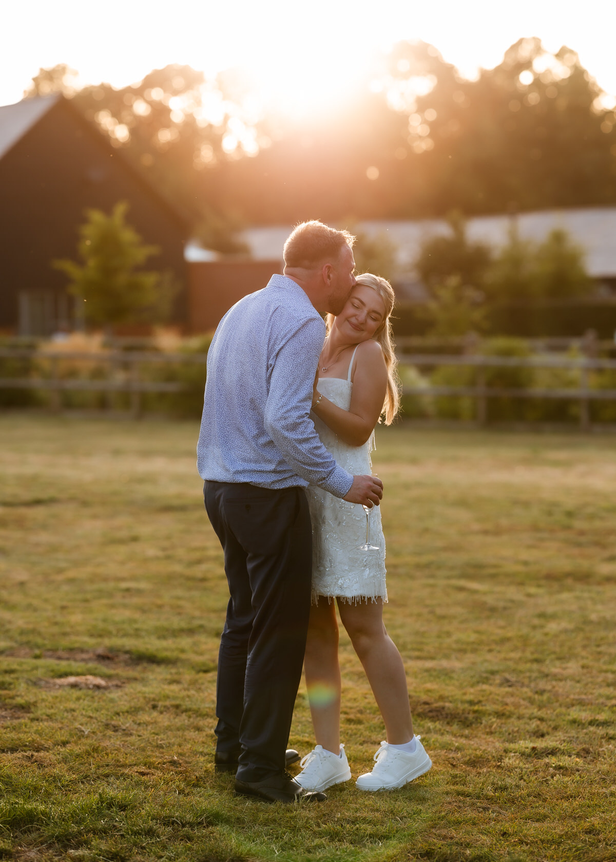 A man kisses a smiling woman on the temple as they stand in a sunny grassy field. She wears a white dress and sneakers, while he holds a drink. The sun sets behind them, casting a warm glow.