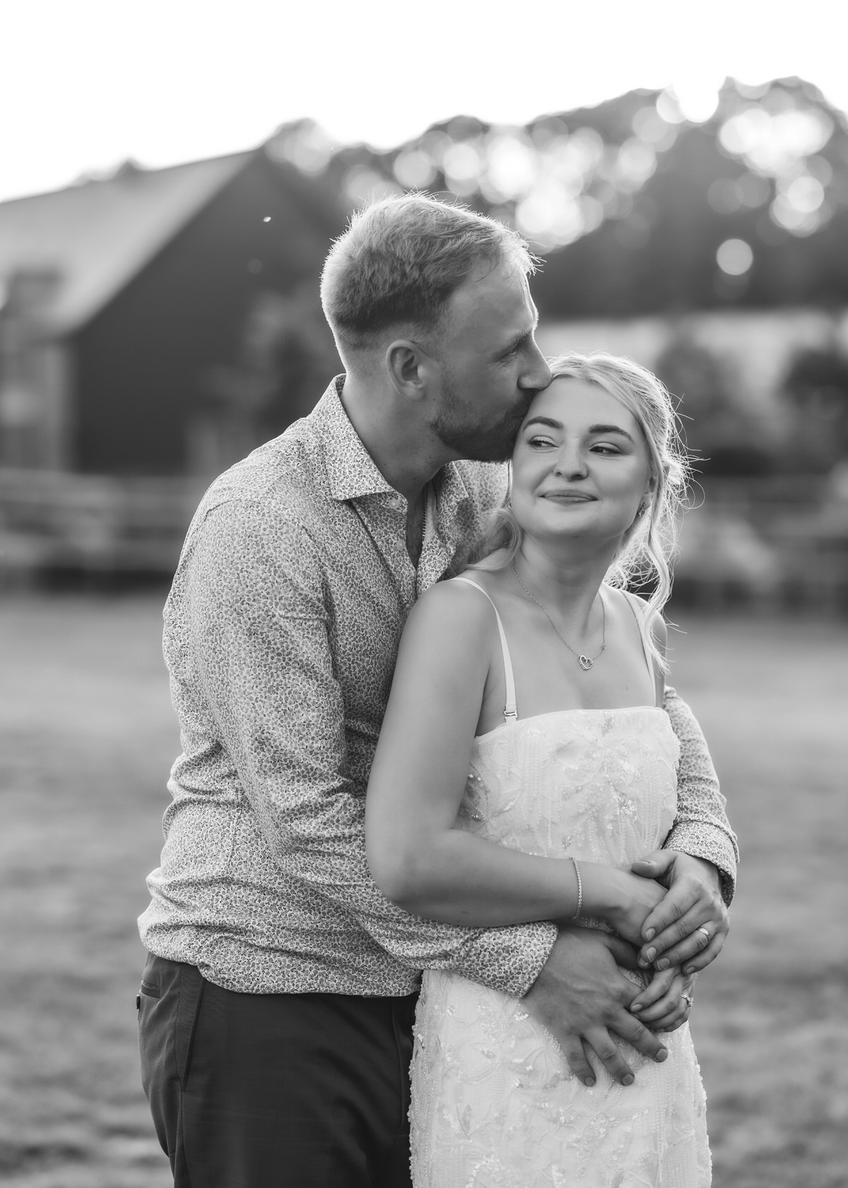 A man hugs a smiling woman from behind and kisses her forehead outside. Both appear happy. The image is in black and white, with a blurred background of trees and buildings.