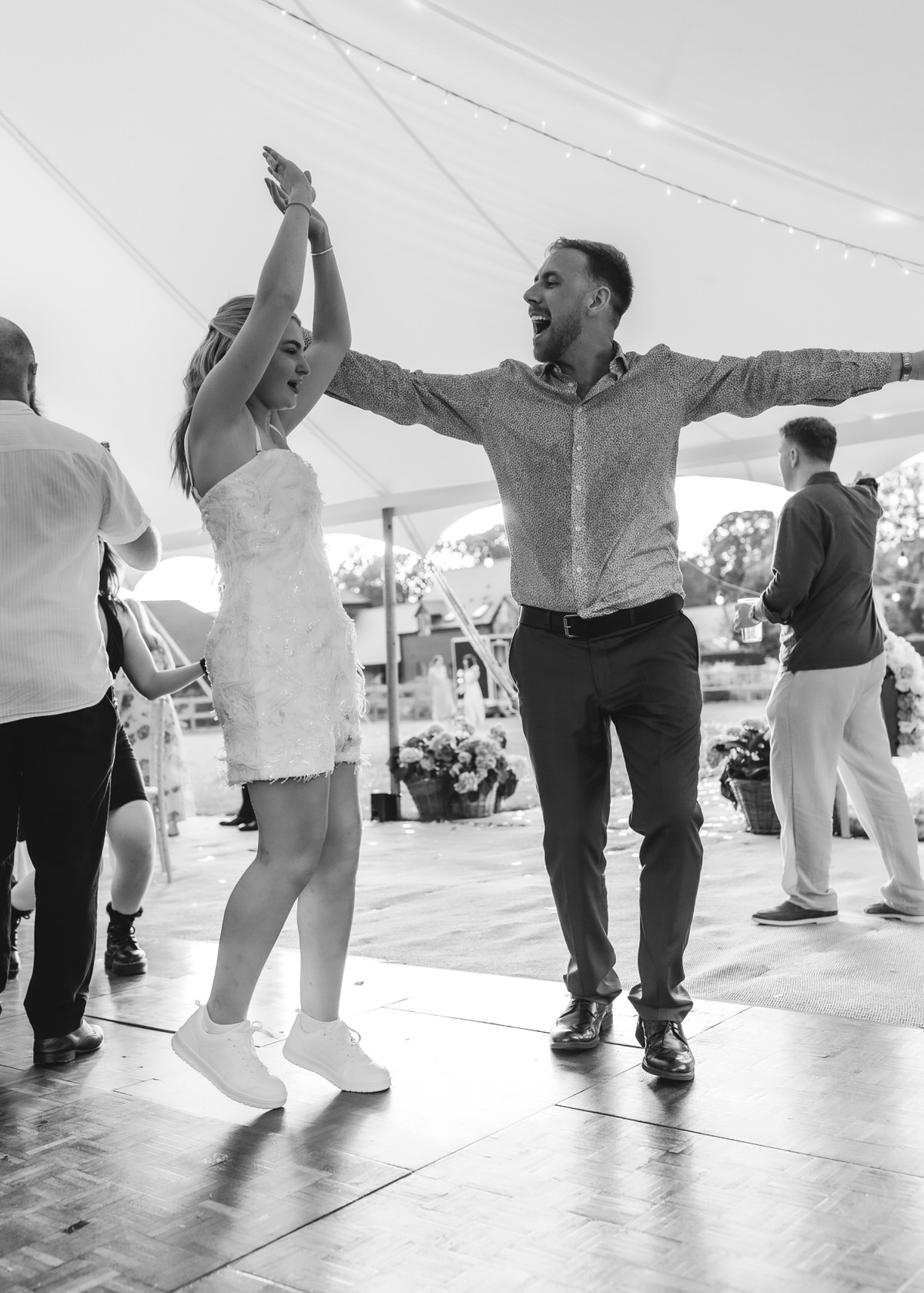 A woman in a short dress and sneakers dances with a man in a button-up shirt under a tent at an event. Both have their arms raised and look joyful. Other people and decorations are visible in the background.