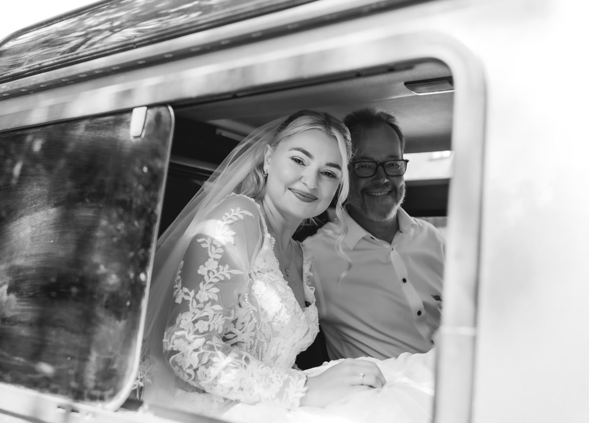 A smiling bride in a lace dress and a man in glasses sit together inside a car, visible through the open window. The photo is black and white.