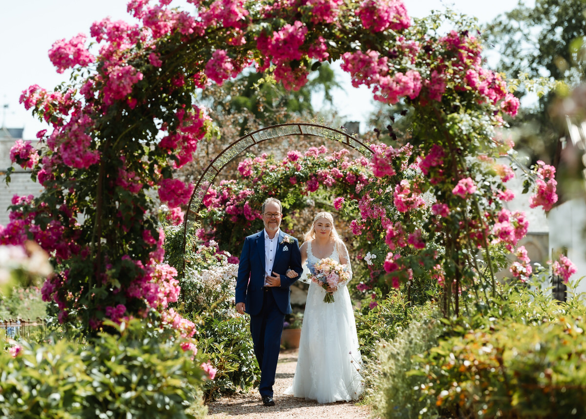 A bride in a white dress holding a bouquet walks arm-in-arm with a man in a blue suit under pink flower arches in a sunny garden.