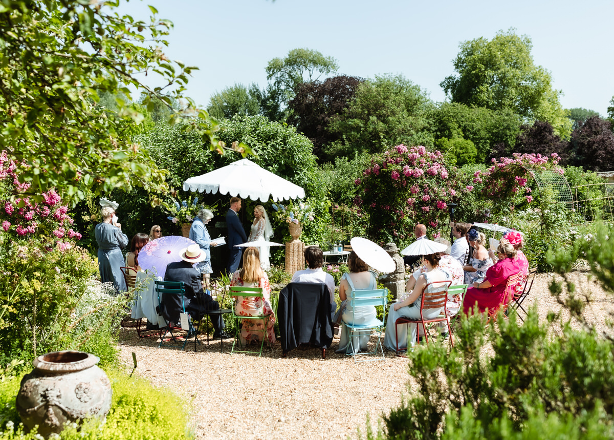 A group of people in colorful outfits and hats are seated outdoors in a lush garden, attending an event under a small white canopy on a sunny day.