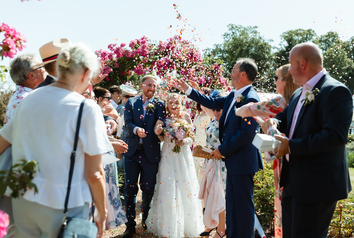 A bride and groom walk outdoors, smiling, as guests toss confetti in celebration. The scene is bright, colorful, and joyful, with flowers and greenery in the background.