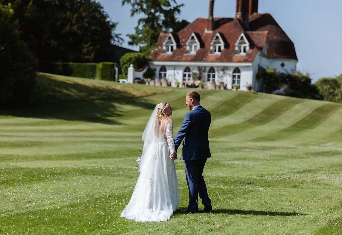 A bride and groom stand hand in hand on a green lawn, facing each other, with a large, elegant house and trees in the background under a sunny sky.