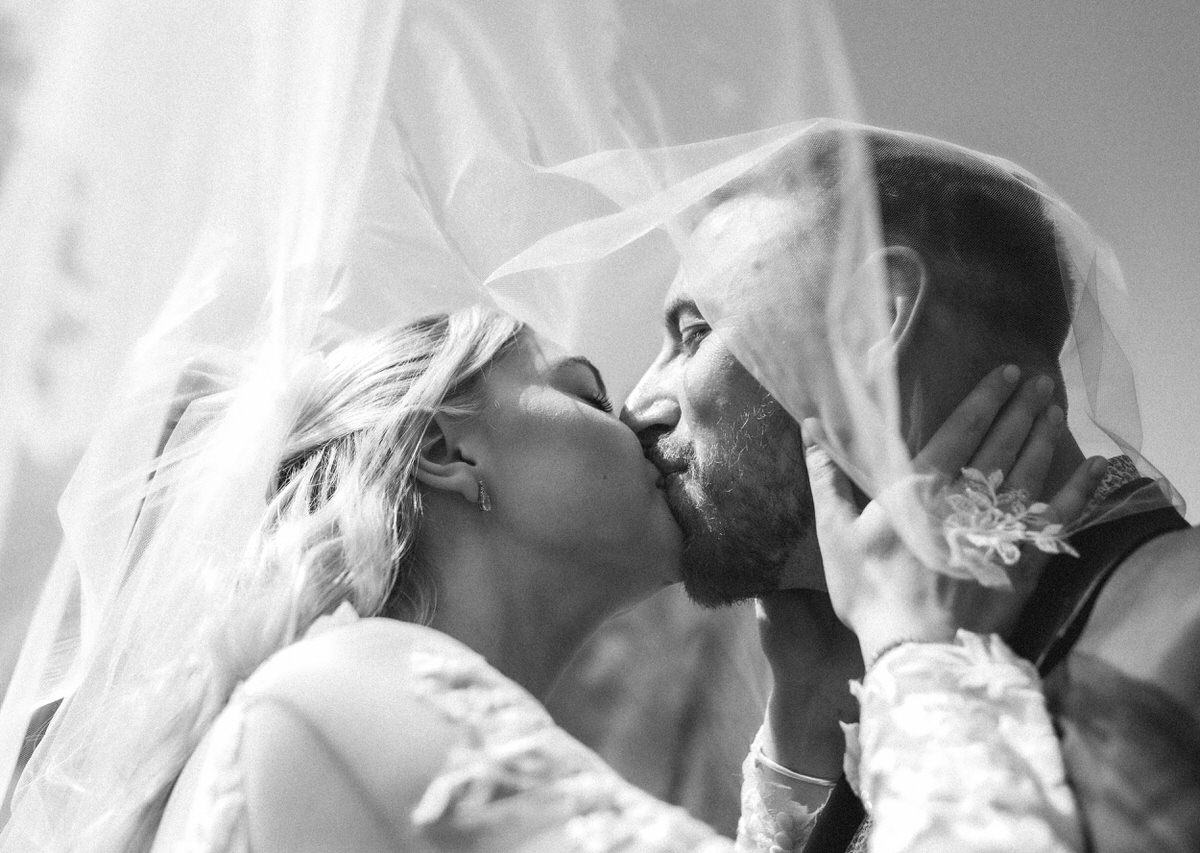 A bride and groom share a kiss under a flowing veil on their wedding day, with sunlight softly illuminating their faces in a romantic, close-up black and white photo.