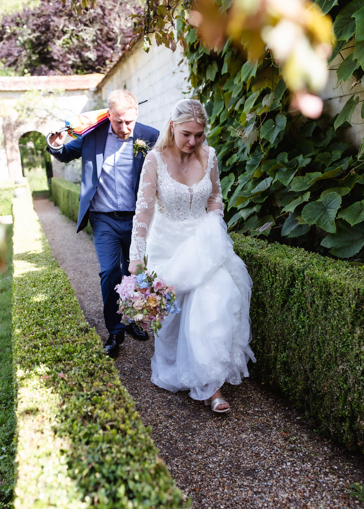 A bride in a white lace dress holds a bouquet and walks along a garden path, followed by a man in a suit draping a colorful scarf over his shoulder. Lush green foliage lines the walkway.