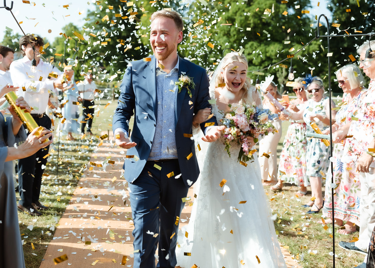 A smiling bride and groom walk arm-in-arm as guests throw gold confetti during an outdoor wedding celebration. Guests cheer and take photos in the background under sunny skies.