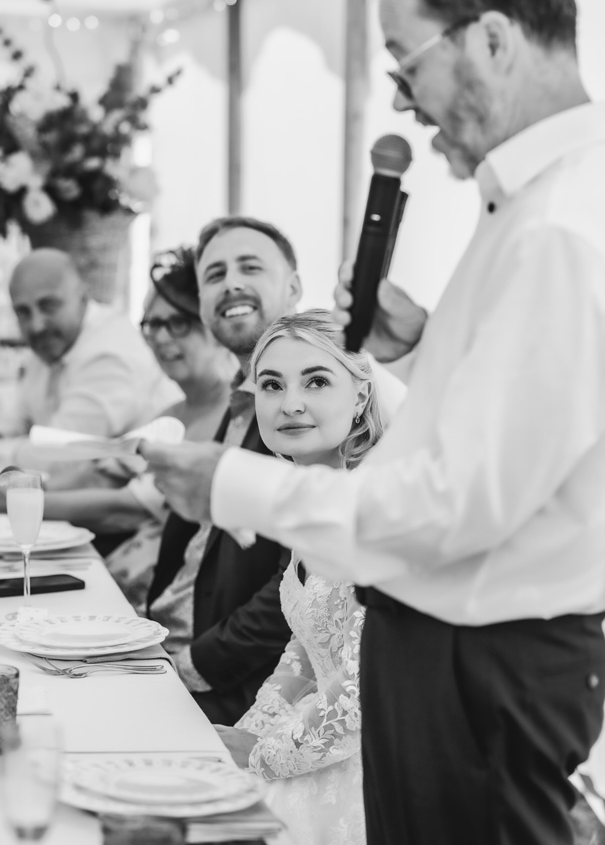A bride in a lace dress sits at a table, smiling up at a man giving a speech with a microphone. Other guests, including a man and woman, smile and listen. The setting appears to be a wedding reception.