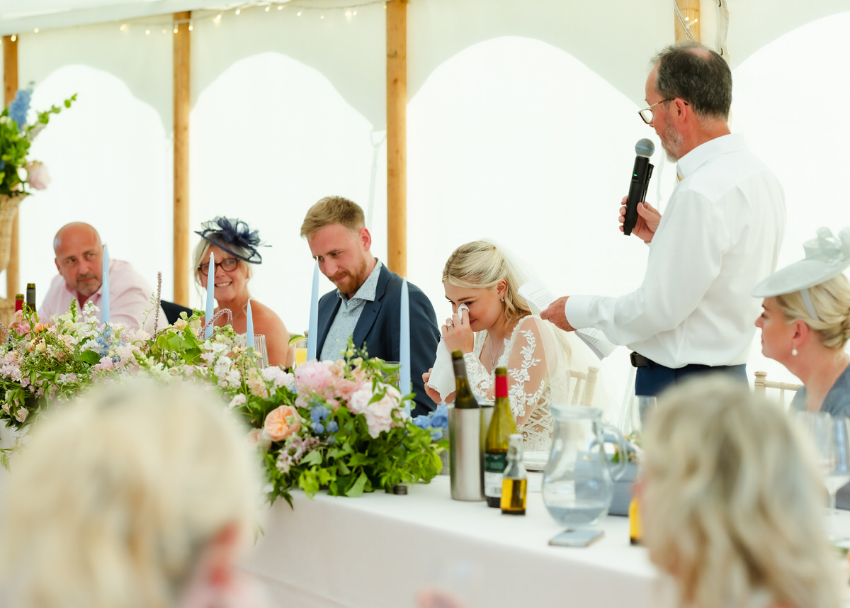 A bride wipes away tears while listening to a speech at a wedding reception. Other guests at the table smile and listen. The table is decorated with flowers and drinks.