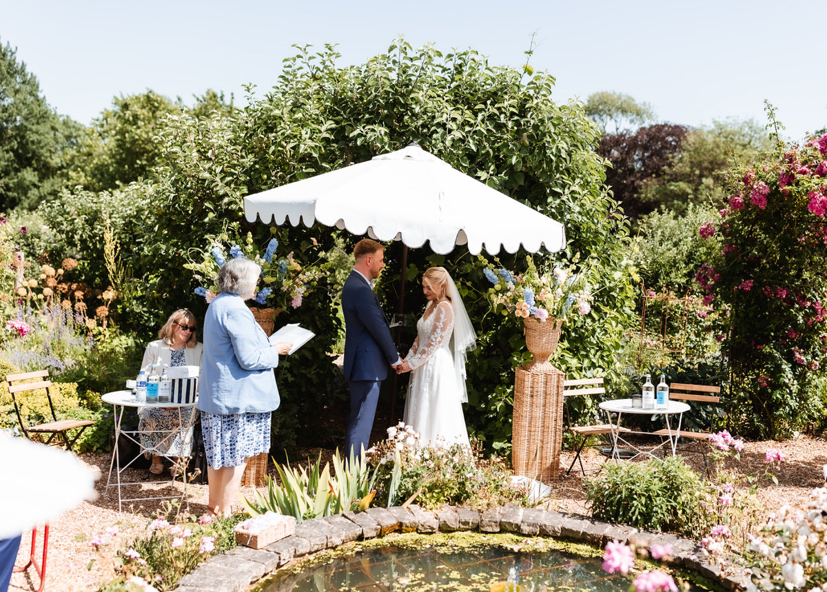 A bride and groom stand under a white umbrella in a garden exchanging vows, with an officiant and two seated guests nearby, surrounded by greenery and flowers next to a small pond.