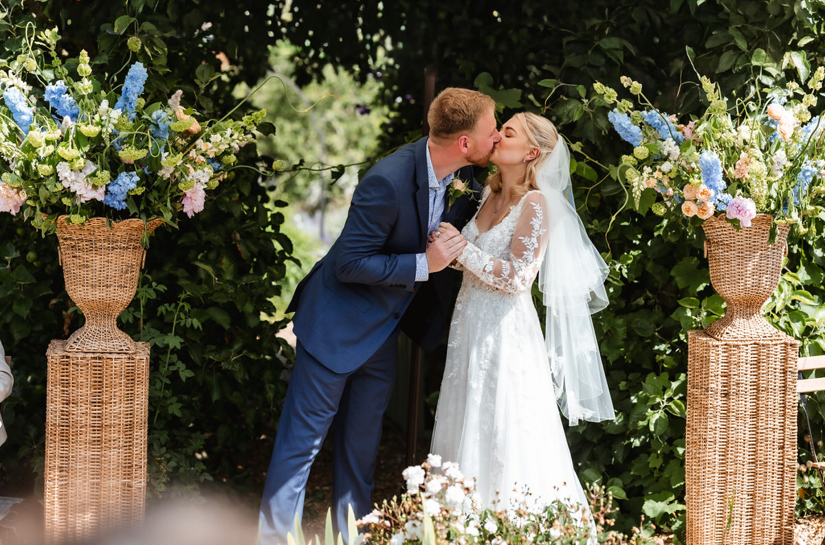 A bride and groom share a kiss at their outdoor wedding ceremony, standing between two large wicker vases filled with colorful flowers and greenery.