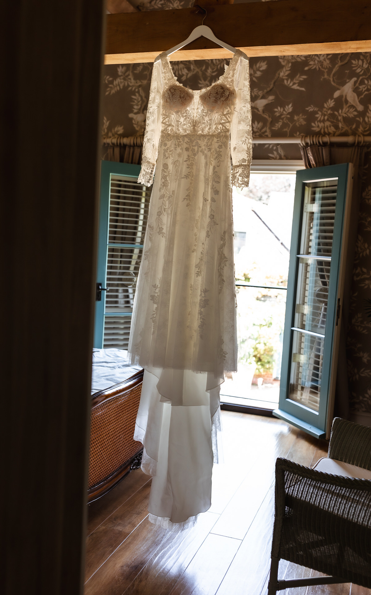 A long white lace wedding dress hangs on a hanger in a sunlit room with open blue doors, wooden floors, and floral wallpaper.