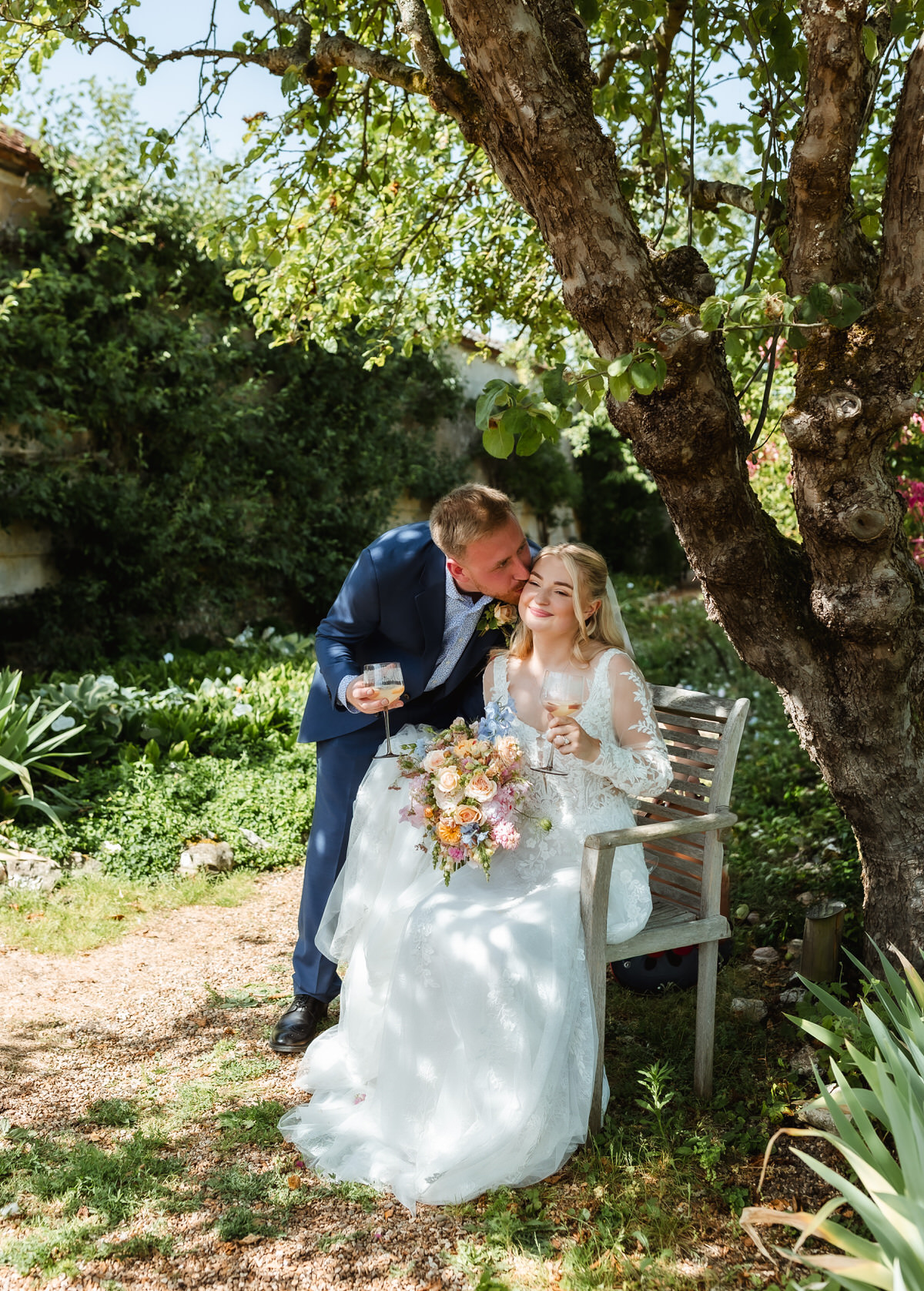 A groom in a blue suit kisses the bride on the cheek as she sits on a bench holding a bouquet, both smiling under a tree in a sunlit garden.