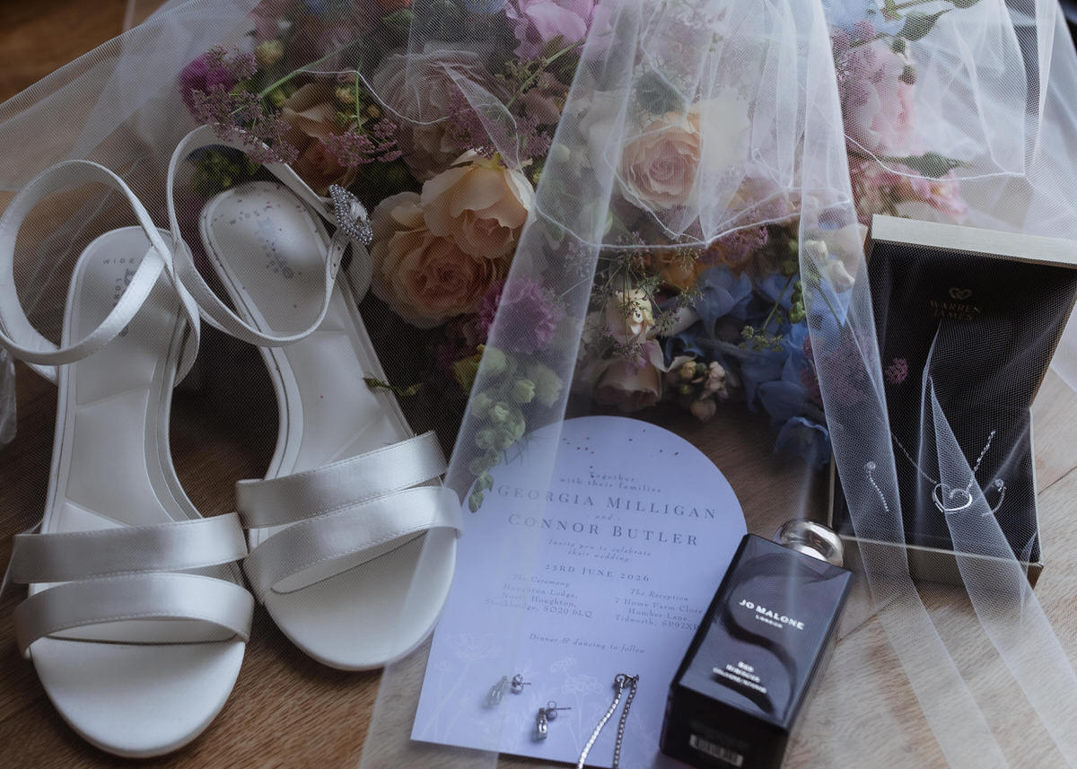 White bridal shoes, floral bouquet, wedding invitation, Jo Malone perfume, earrings, necklace, and veil arranged on a wooden surface, creating an elegant wedding flat lay.