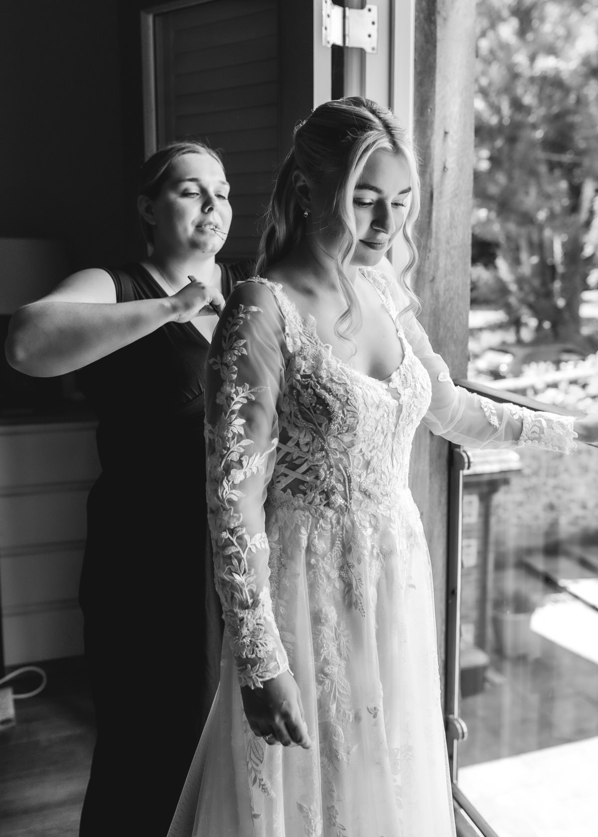 A woman in a wedding dress stands near a window while another woman helps adjust the back of her gown. Natural light streams in, highlighting the bride’s lace sleeves. Both women appear focused on the preparation.