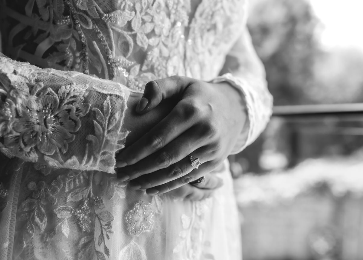 A close-up, black and white photo of a person in an embroidered lace dress, with hands gently clasped at the waist, showcasing a ring on one finger.