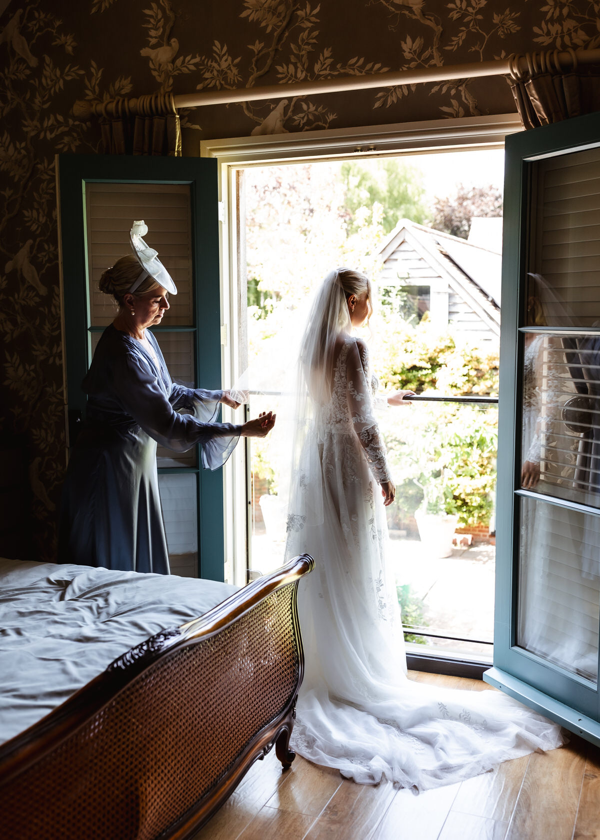 A bride in a white dress and veil stands by open doors, looking outside. An older woman in a formal outfit helps adjust her veil. Sunlight shines in from the garden.