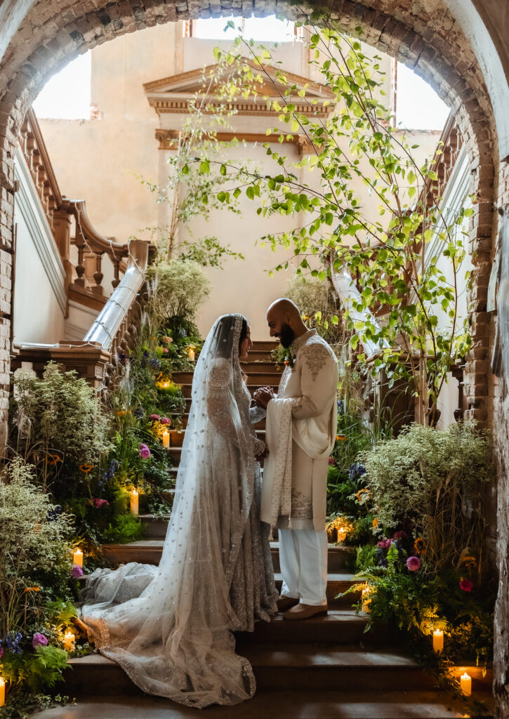 A bride and groom stand together on a flower-lined staircase, holding hands and facing each other in a romantic, historic setting filled with greenery and soft candlelight—captured beautifully by a Hampshire wedding photographer.
