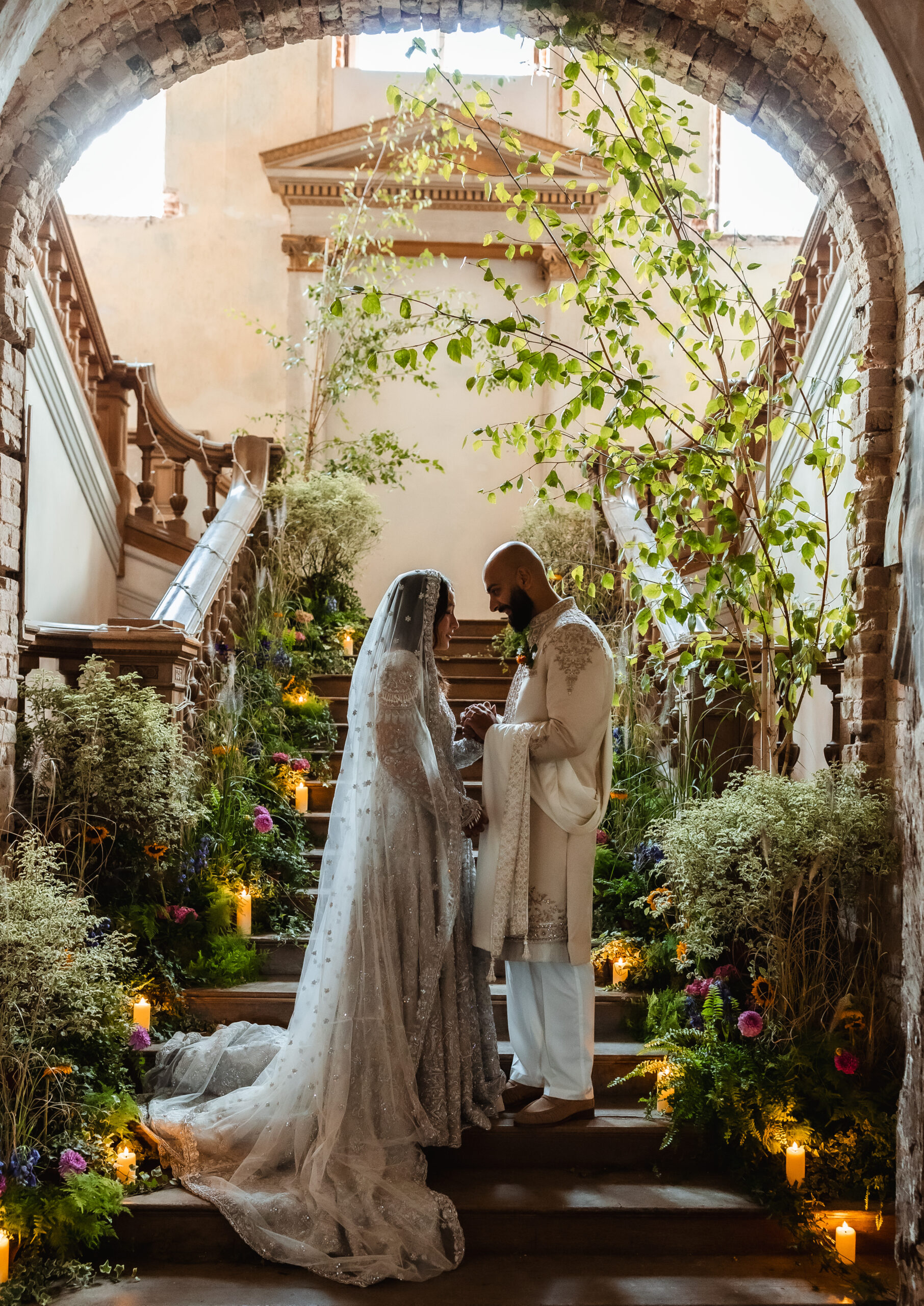 A bride and groom stand together on a flower-lined staircase, holding hands and facing each other in a romantic, historic setting filled with greenery and soft candlelight—captured beautifully by a Hampshire wedding photographer.