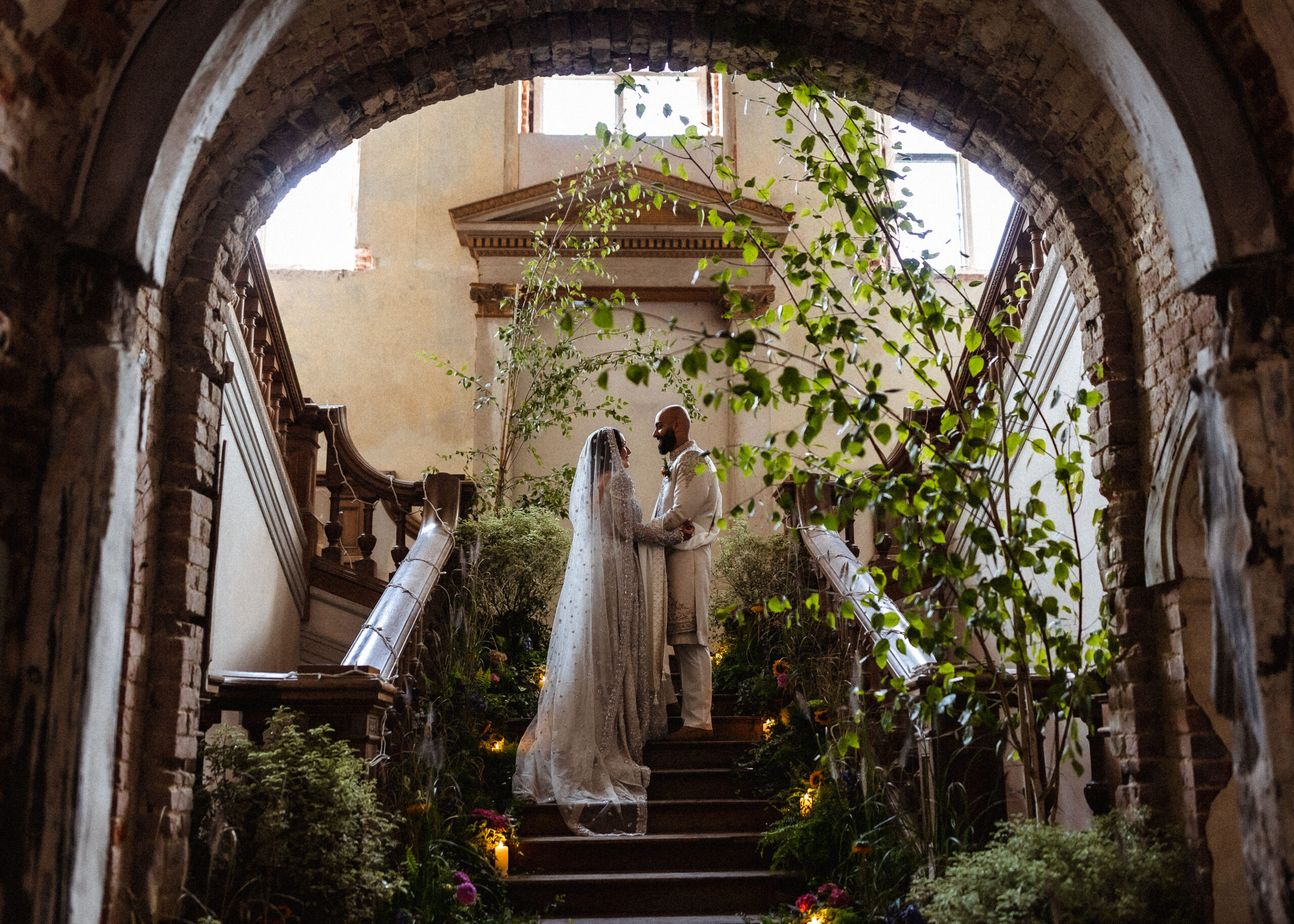 A couple in wedding attire stands on a stone staircase, surrounded by greenery and flowers, under an arched, rustic ceiling in a softly lit, historic building—captured beautifully by a Hampshire wedding photographer.