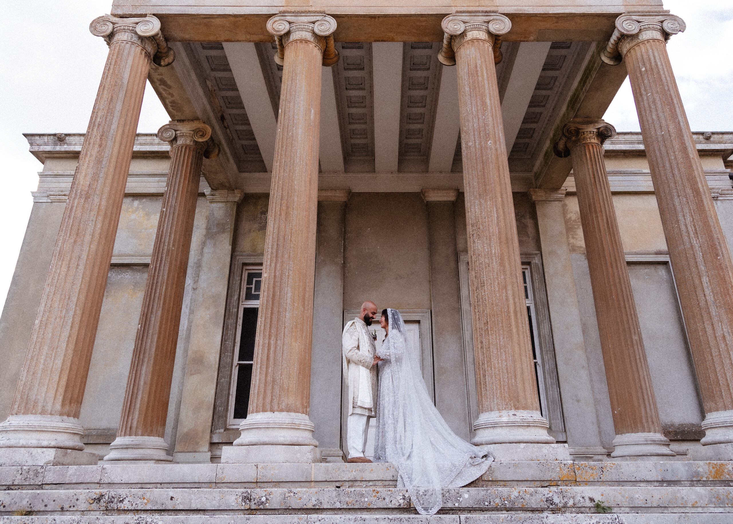 A couple in wedding attire stands on stone steps, embracing beneath tall, classical columns of an old, grand building—beautifully captured by a Hampshire wedding photographer.