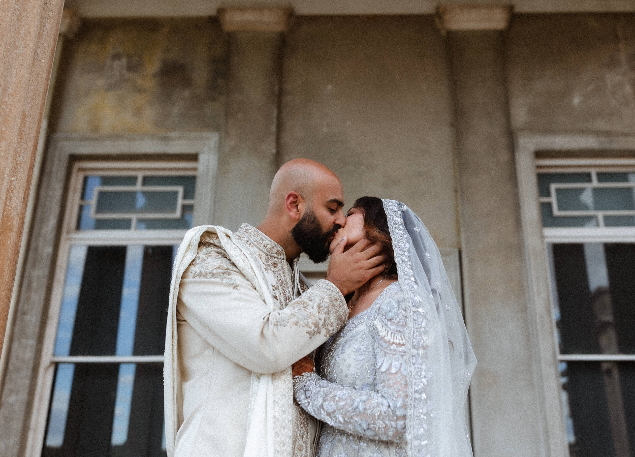 A couple dressed in traditional wedding attire shares a gentle kiss in front of a weathered building with large windows, beautifully captured by a Hampshire wedding photographer.