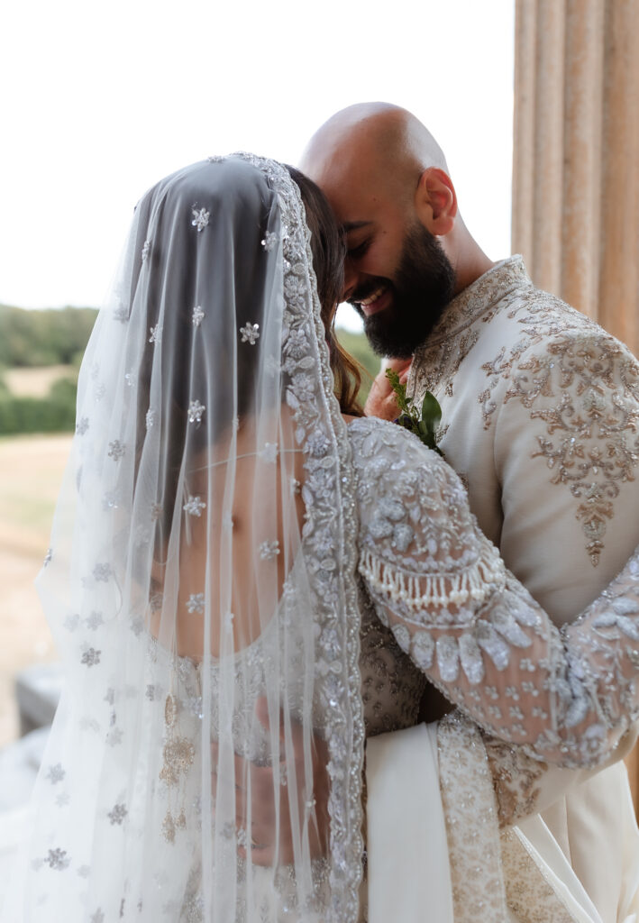 A bride and groom in ornate, traditional attire embrace closely, smiling. The bride’s jeweled veil drapes over her back, while the groom holds her, resting his forehead gently against hers—a timeless moment beautifully captured by a Hampshire wedding photographer.
