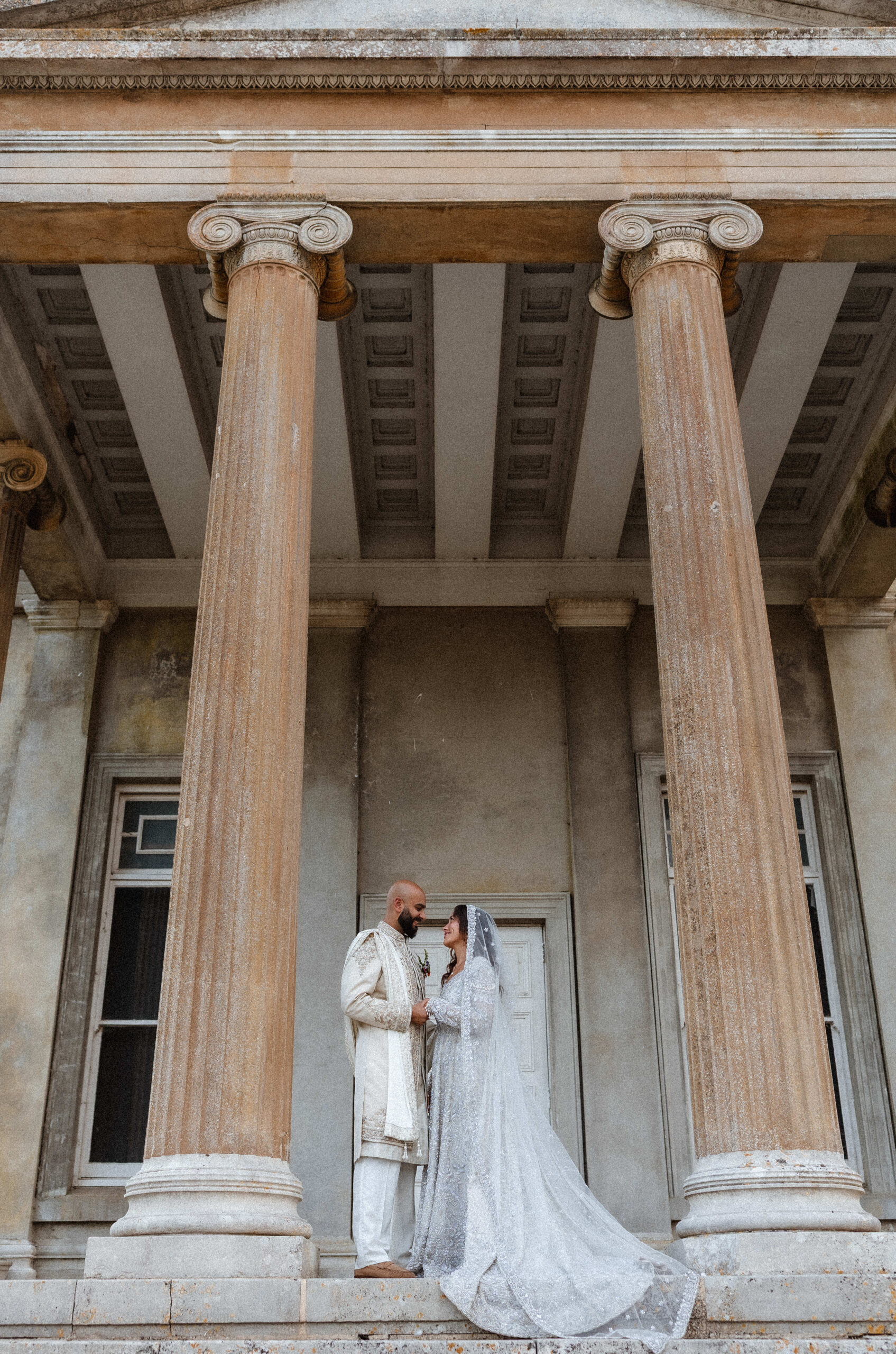 A couple in traditional wedding attire stands on steps between large stone columns, gazing at each other in front of an old, grand building—captured beautifully by a Hampshire wedding photographer.