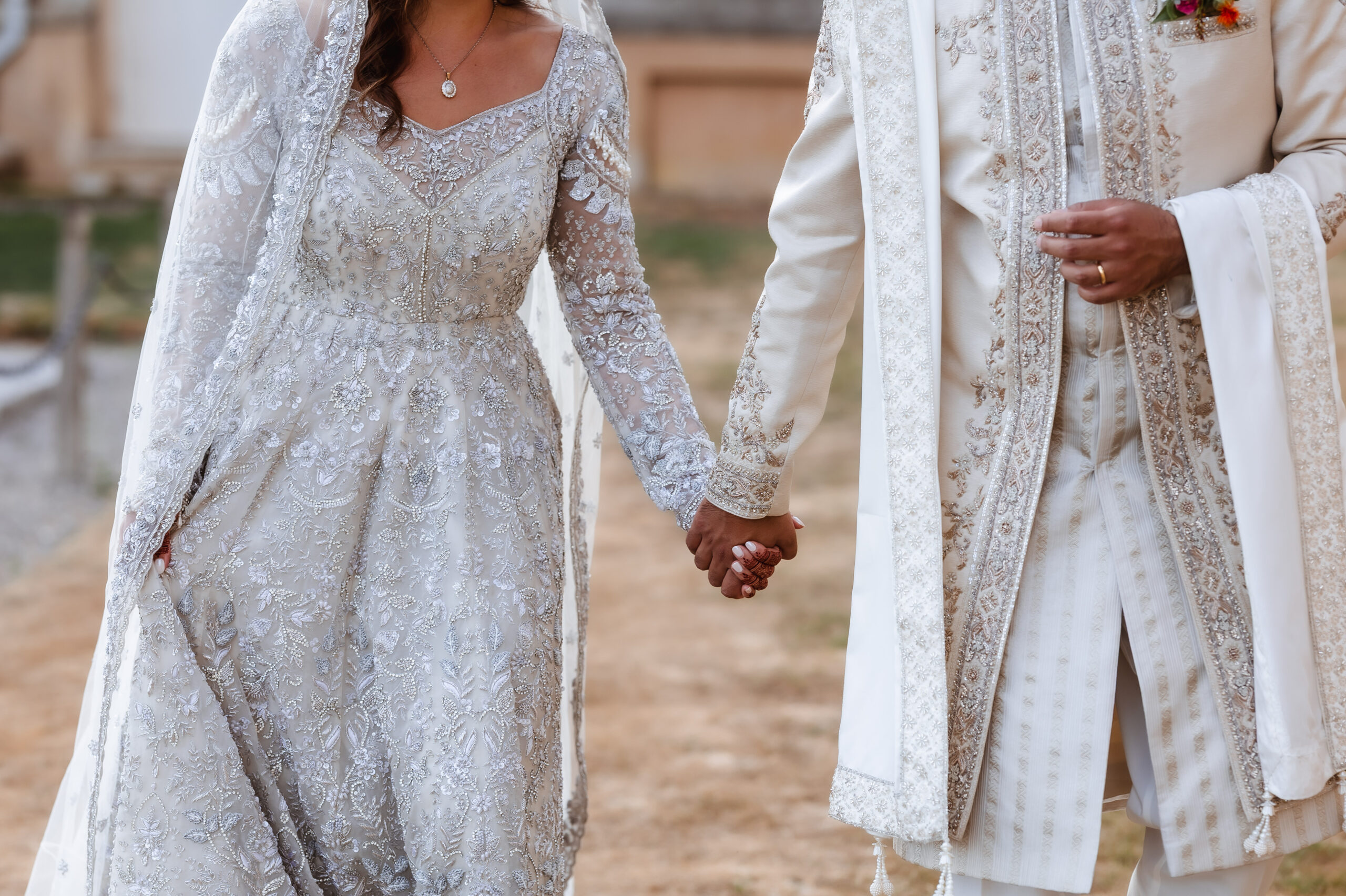 A bride and groom in ornate, light-colored traditional attire hold hands outdoors, showcasing detailed embroidery on their wedding outfits, all beautifully captured by a Hampshire wedding photographer.