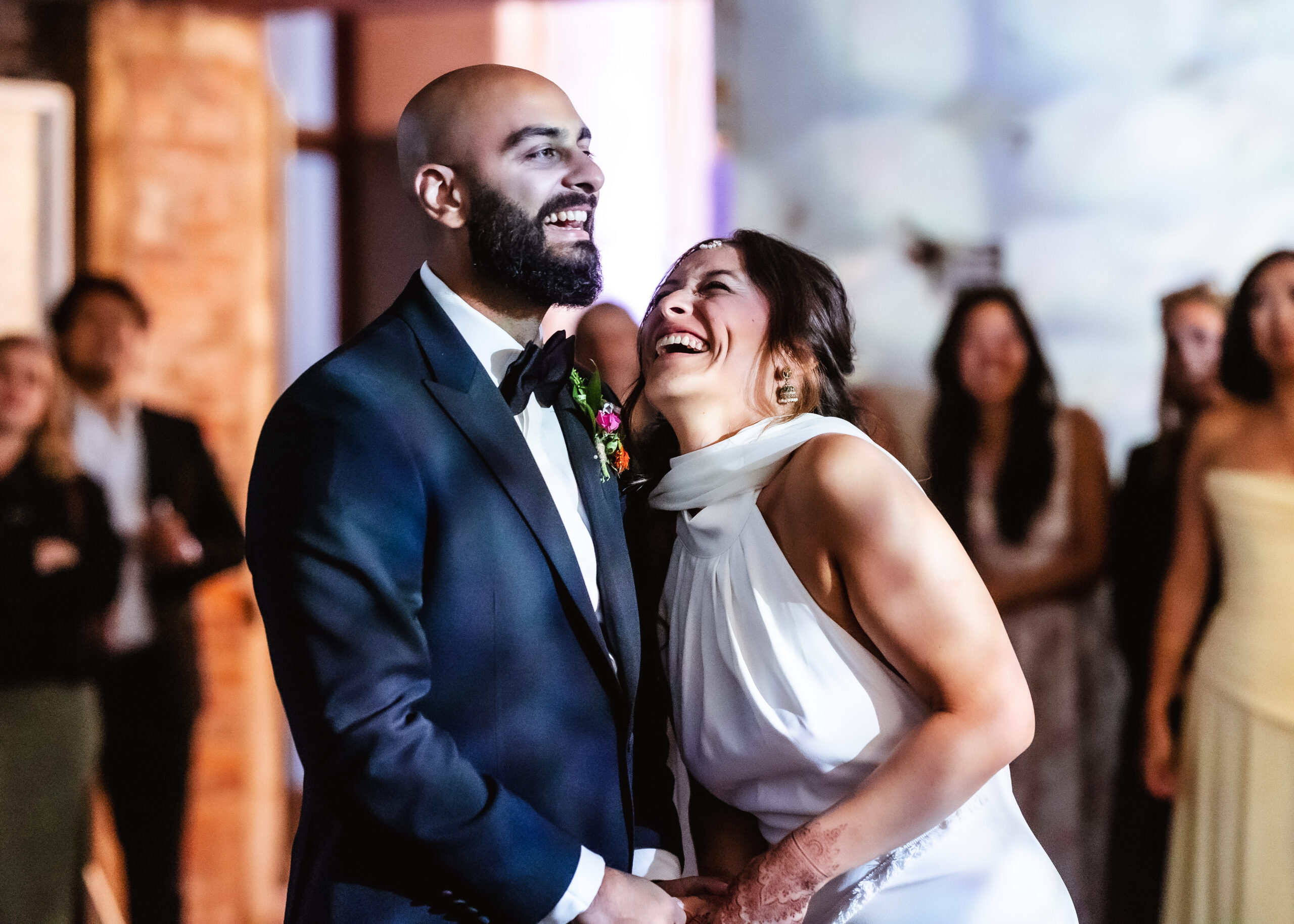 A newlywed couple, dressed in formal attire, laugh joyfully while holding hands on the dance floor. Guests in the background smile and watch the couple, bathed in warm, festive lighting—a perfect moment beautifully captured by a Hampshire wedding photographer.