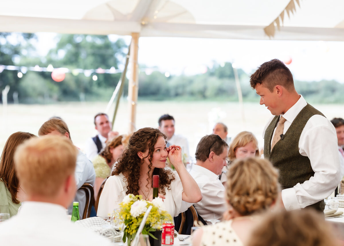 A woman with curly hair wipes her eye while a man in a vest speaks to her. They are at a wedding reception under a tent, surrounded by guests seated at decorated tables—beautifully captured by a country-house wedding photographer.