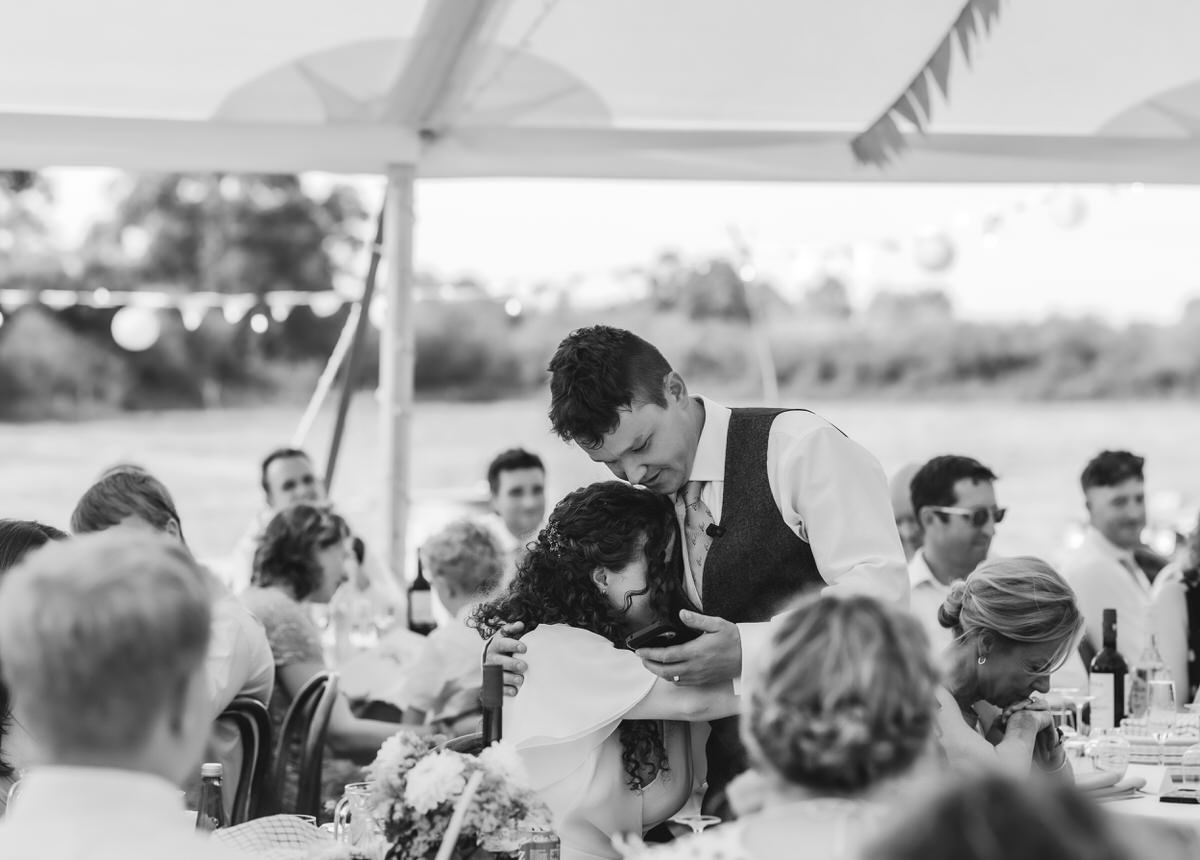 A black-and-white photo by a country-house wedding photographer captures a couple embracing and smiling at their wedding reception, surrounded by seated guests under a tent decorated with bunting.
