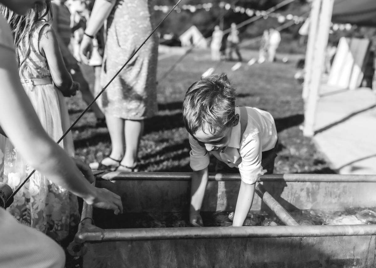 A young boy leans over a metal tub of water, reaching in with both hands. Other children and adults stand nearby on a grassy area during an outdoor event, reminiscent of candid moments captured by a country-house wedding photographer. The image is black and white.
