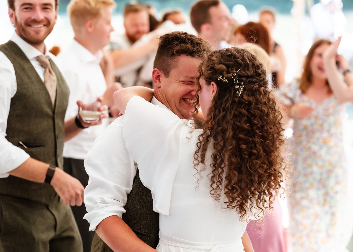 A joyful couple embraces and smiles while dancing at a lively wedding reception, surrounded by guests who are clapping and celebrating, perfectly captured by a country-house wedding photographer.