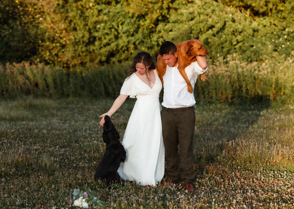A woman in a white dress and a man in a white shirt stand in a field with two dogs, one black standing near the woman and one brown draped over the man’s shoulders—a scene beautifully captured by a country-house wedding photographer.
