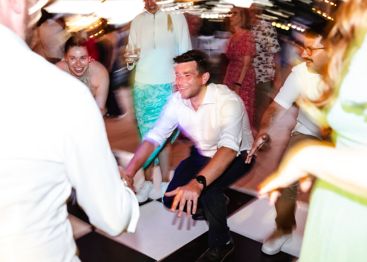 A group of people dance energetically on a checkered floor at a lively event, as vibrant lights and blurred motion convey excitement and joy—capturing the perfect moment for any country-house wedding photographer.