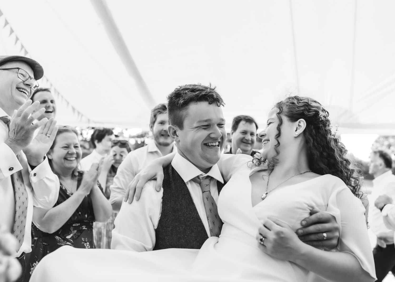 A groom smiles while carrying his laughing bride as friends and family cheer and clap around them at a joyful wedding celebration under a tent, beautifully captured by a country-house wedding photographer.