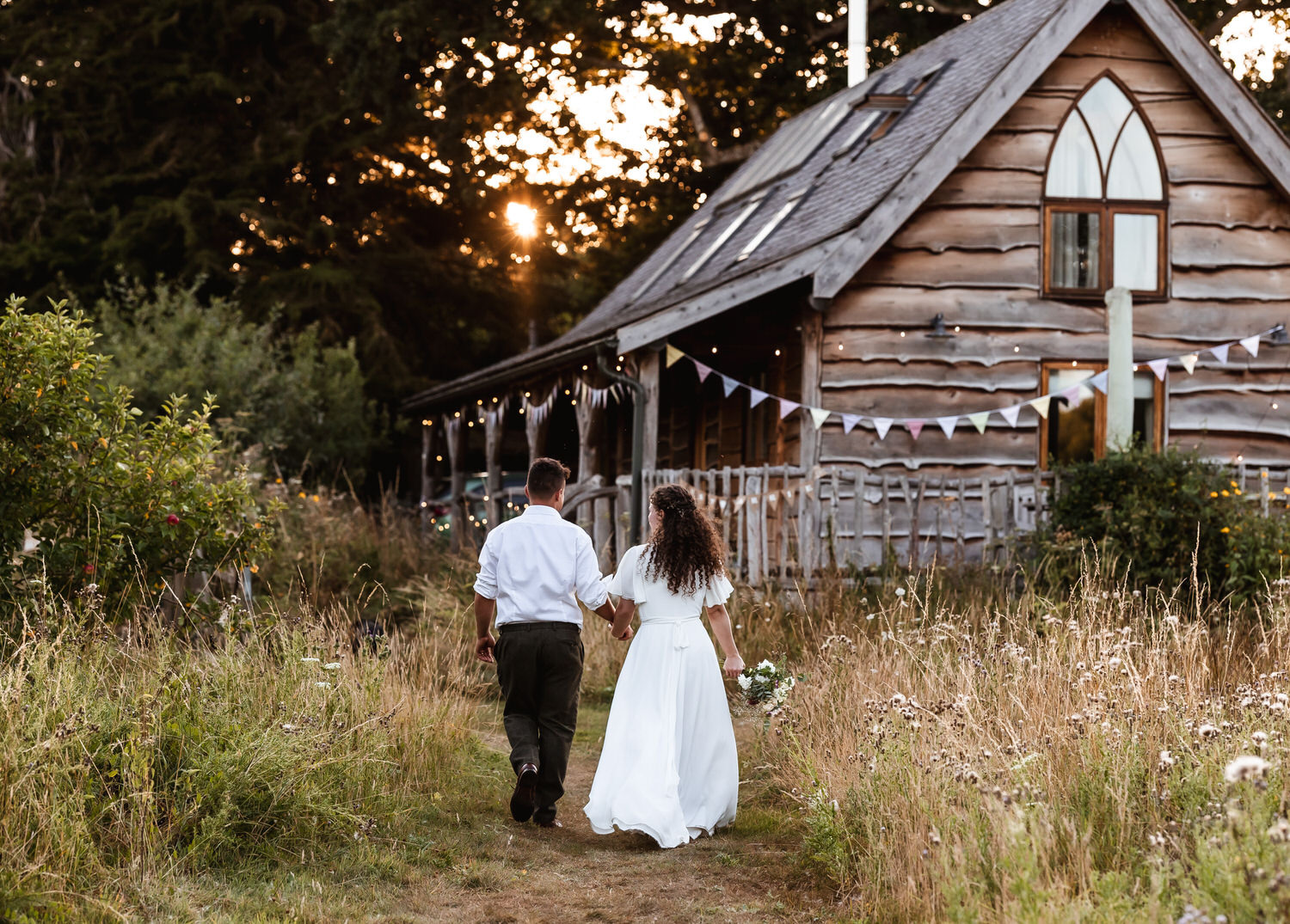 A couple walks hand in hand toward a rustic wooden cabin surrounded by tall grass and wildflowers at sunset, with bunting decorating the porch—an idyllic setting captured perfectly by a country-house wedding photographer.