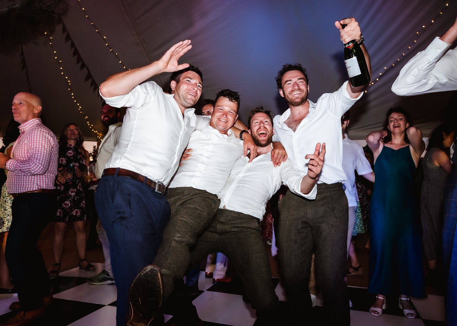 Four men in white shirts and dark pants celebrate on a dance floor, smiling and posing energetically; one holds a wine bottle. Captured by a country-house wedding photographer, the lively scene unfolds against a festive background of people and glowing string lights.