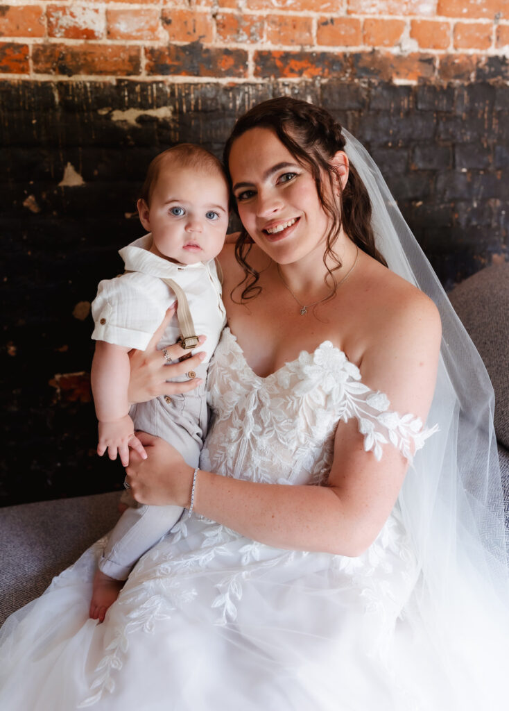 A bride in a white lace dress and veil sits, smiling, while holding a baby boy wearing suspenders on her lap. They are posed in front of a rustic brick and black wall.