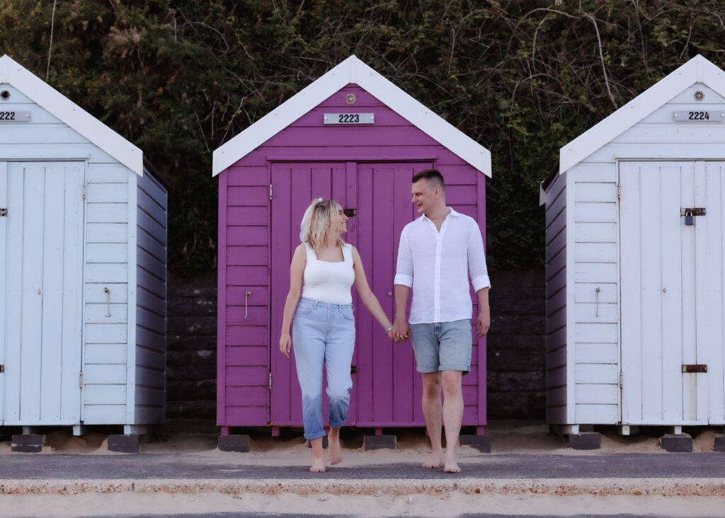 A couple holding hands and smiling at each other stands barefoot in front of a purple beach hut, flanked by two white huts, with greenery in the background.