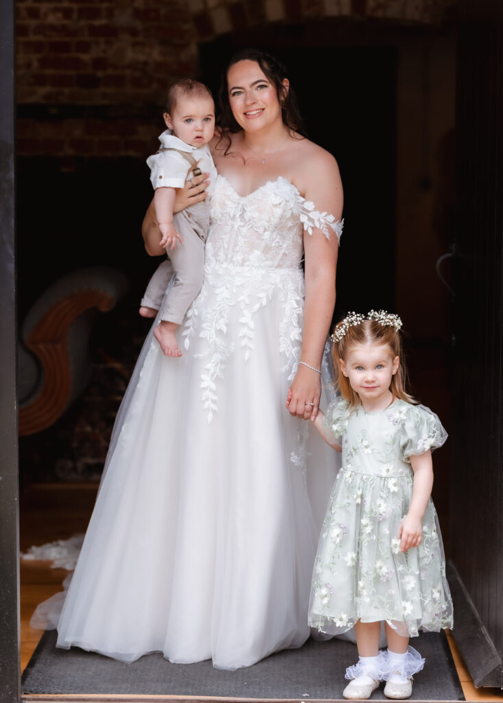 A smiling bride in a white dress stands holding a baby boy, with a little girl in a light green dress and flower crown standing beside her.