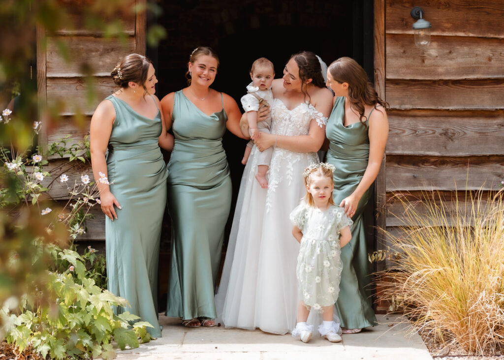 A bride in a white dress stands with four bridesmaids in green dresses and two young children outside a rustic wooden building, all smiling and posing together.