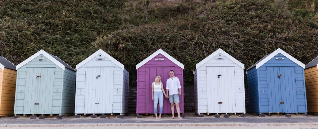 A woman and man stand in front of a purple beach hut, surrounded by four other colorful huts with green foliage behind them.