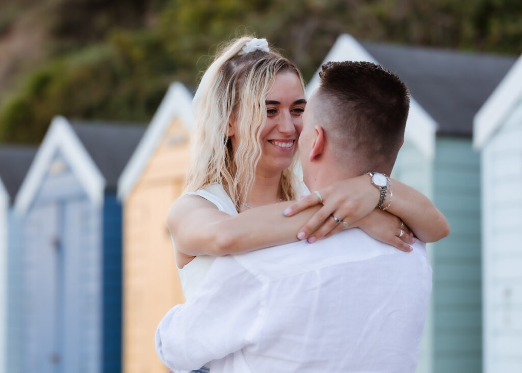 A woman smiles and embraces a man outdoors, with colorful beach huts in the background.