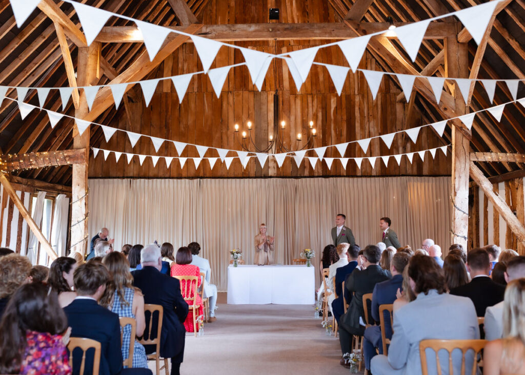 Rows of people seated in a rustic barn with wooden beams, white bunting overhead, and a ceremony at the front featuring an officiant and two people standing by a white table.