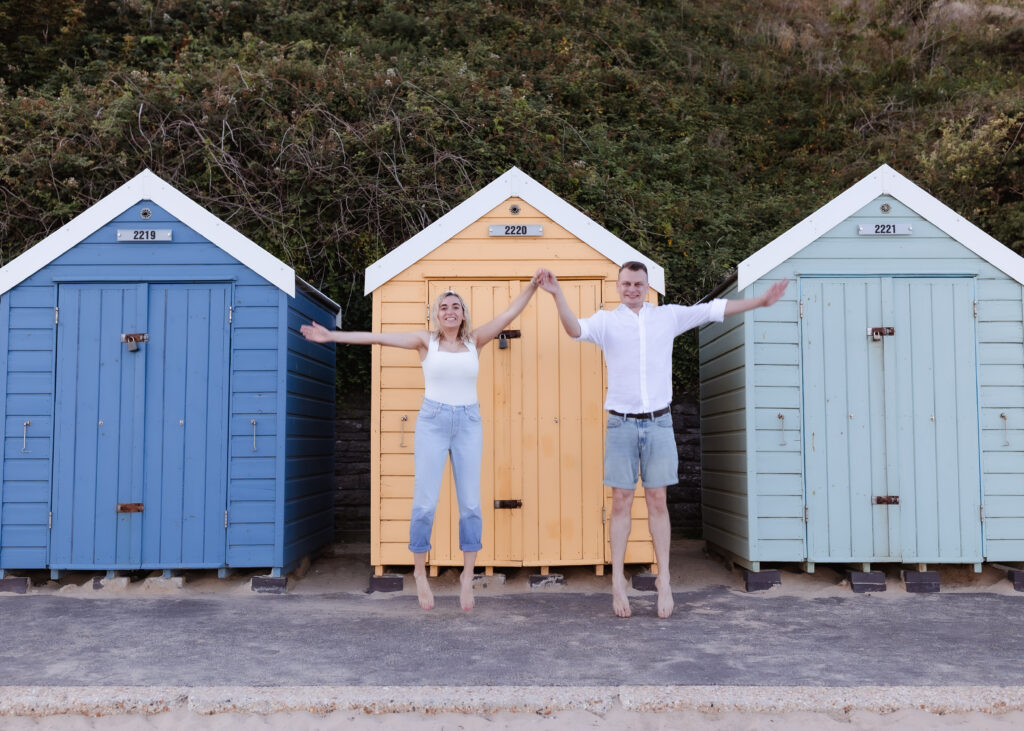 A smiling couple holds hands and jumps in front of three colorful beach huts—blue, yellow, and light blue—set against a backdrop of greenery.