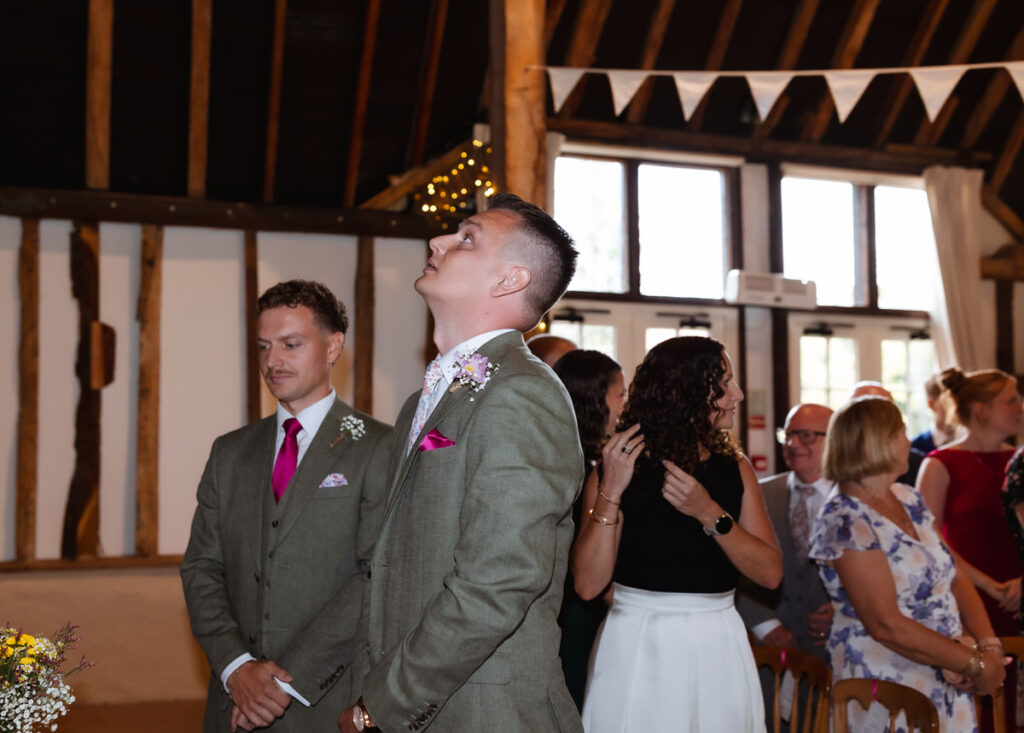 A man in a green suit looks up with anticipation, standing beside another man at a wedding ceremony in a rustic venue with wooden beams and guests seated in the background.